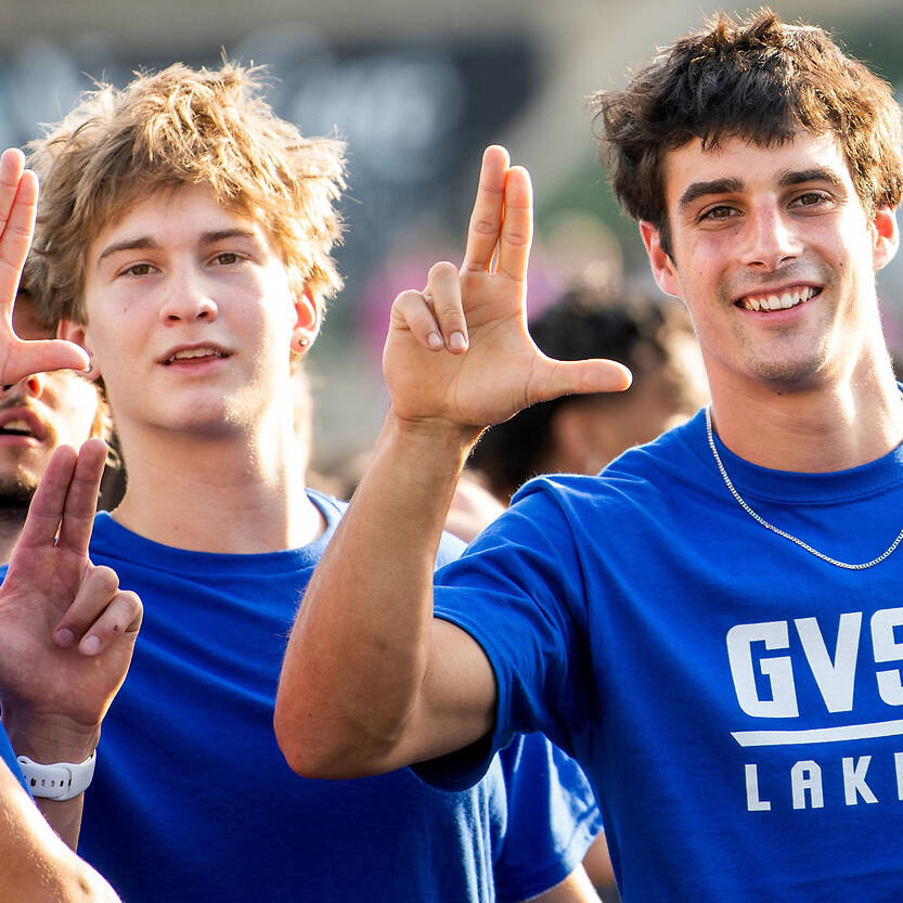Three individuals in blue GVSU Lakers shirts make peace signs while smiling at the camera.